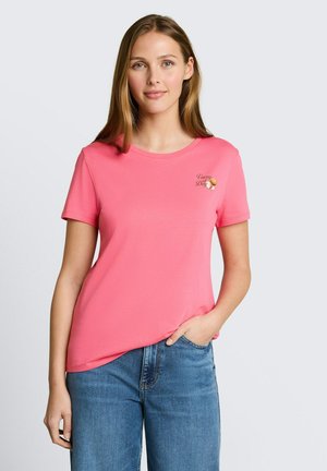 Kobieta w różowym t-shircie z krótkim rękawem z napisem "Vacay All Day" i grafiką kokosa, w połączeniu z niebieskimi dżinsami, stoi na jednolitym tle.