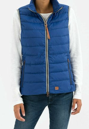 Persoon draagt een blauw gewatteerd mouwloos vest over een wit shirt met lange mouwen en donkerblauwe spijkerbroek, staat met handen ontspannen langs het lichaam.