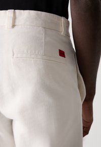 Hvit kanvasbukse med en baklomme som har en rød "Hugo"-etikett. Stoffet har en teksturert overflate og en tilpasset silhuett.
