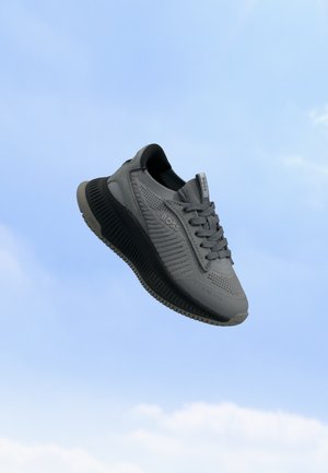 Baskets BOSS grises et noires avec texture tricotée et lacets, flottant sur un fond de ciel bleu parsemé de légers nuages.