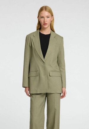 Blazer verde oliva con maniche lunghe, colletto a revers e due tasche frontali, abbinato a pantaloni coordinati. Tessuto morbido e lavorato.