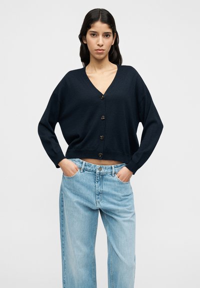 Καζάκα ριγωτή σε ναυτικό μπλε χρώμα από μαλακό πλεκτό υλικό με V-λαιμόκοψη, διαθέτει πέντε κουμπιά στο μπροστινό μέρος και φαρδιά μακριά μανίκια, συνδυασμένη με ανοιχτόχρωμο μπλε τζιν.