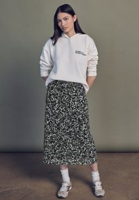 Weißer Kapuzenpullover mit Text, kombiniert mit einem langen schwarzen Rock mit einem weißen Blumenmuster, abgerundet mit grauen Turnschuhen und weißen Socken.