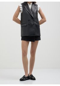 Svart läder väst utan ärmar med dubbelknäppta knappar, slät tekstur och fickor med klaff, i kombination med en svart denimkjol och svarta loafers.