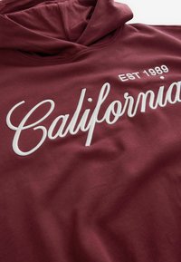 Bordeauxfarvet hættetrøje med en blød tekstur. Har hvid broderet "California" -tekst og "EST 1989" i et rent, afrundet skrifttype.