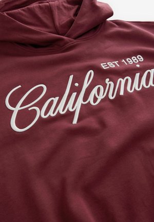 Bordeauxfarvet hættetrøje med en blød tekstur. Har hvid broderet "California" -tekst og "EST 1989" i et rent, afrundet skrifttype.