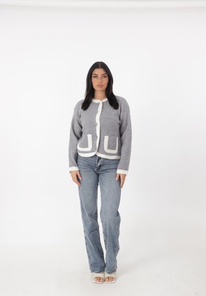 Grijze gebreide cardigan met witte accenten, voorzien van een ronde hals, voorzakken en knoopsluiting. Gecombineerd met lichtblauwe jeans.