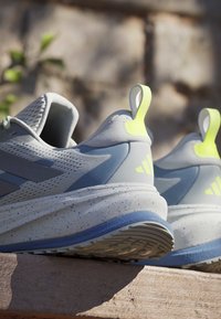 Chaussures de course gris clair et bleu avec des semelles intermédiaires mouchetées et des languettes de talon jaune néon, placées à l'extérieur sur une surface en bois au soleil.