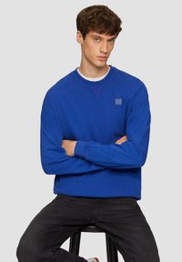Felpa blu con scollo rotondo, polsini a costine e una piccola patch con logo sul petto. Indossata con jeans neri, il modello è seduto.