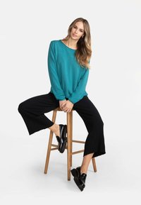 Teal Pullover mit rundem Ausschnitt, kombiniert mit schwarzen, gerippten weit geschnittenen Hosen. Schwarze Schuhe mit glatter Oberfläche und dicken Sohlen.
