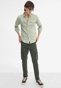 Camisa de botón de color verde claro con dos bolsillos en el pecho, combinada con pantalones cargo verde oscuro que tienen bolsillos laterales y zapatillas negras.