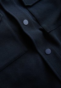 Marineblauer strukturierter Stoff mit einem gerippten Muster, versehen mit zwei hervorgehobenen schwarzen Rundknöpfen und einem Kragen.