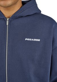 Navy blue zip-up hoodie made of soft fabric, featuring a kangaroo pocket, a metal zipper, and “PEGADOR” logo in white on the left chest.