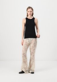 Top noir sans manches associé à un pantalon évasé en dentelle crème transparente avec des motifs floraux ; le modèle se tient contre un fond blanc.