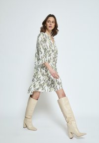 Weißes Kleid mit floralem Muster und grünen Akzenten, lockere Passform, Dreiviertelärmel, kombiniert mit beigefarbenen, knielangen, hohen Stiefeln.