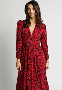 Kobieta z falującymi ciemnymi włosami, ubrana w czarną sukienkę z długim rękawem w czerwony wzór róż, uśmiecha się i stoi na tle jednolitego jasnego tła.