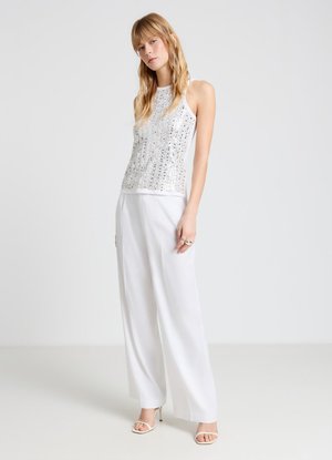 Femme debout portant un top sans manches argenté à paillettes, un pantalon blanc large et des sandales blanches à talons hauts et à bouts ouverts sur un fond uni.