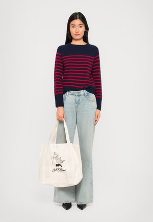 Kvinne står med en marineblå genser med røde striper, lyseblå vide jeans, svarte spisse sko, og holder en hvit tote bag med design av en katt.
