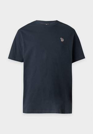 Homme portant un t-shirt noir à manches courtes, un jean large bleu clair et des baskets beige, debout avec une main dans la poche sur fond blanc uni.