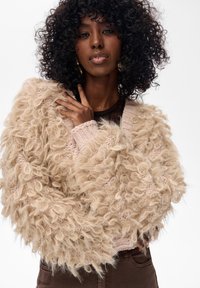 Kobieta z kręconymi czarnymi włosami, nosząca złote kolczyki w kształcie kół i beżowy, fakturowany, puszysty sweter nad ciemnymi brązowymi spodniami.