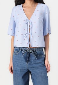 Blouse courte bleu clair avec un profond décolleté en V, manches courtes et lien à nouer à l'avant, présentant un motif rayé et de petites touches florales rouges.