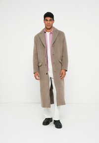 Man som står i en beige långullig kappa, rosa skjorta, vita byxor och svarta skor mot en plain vit bakgrund.