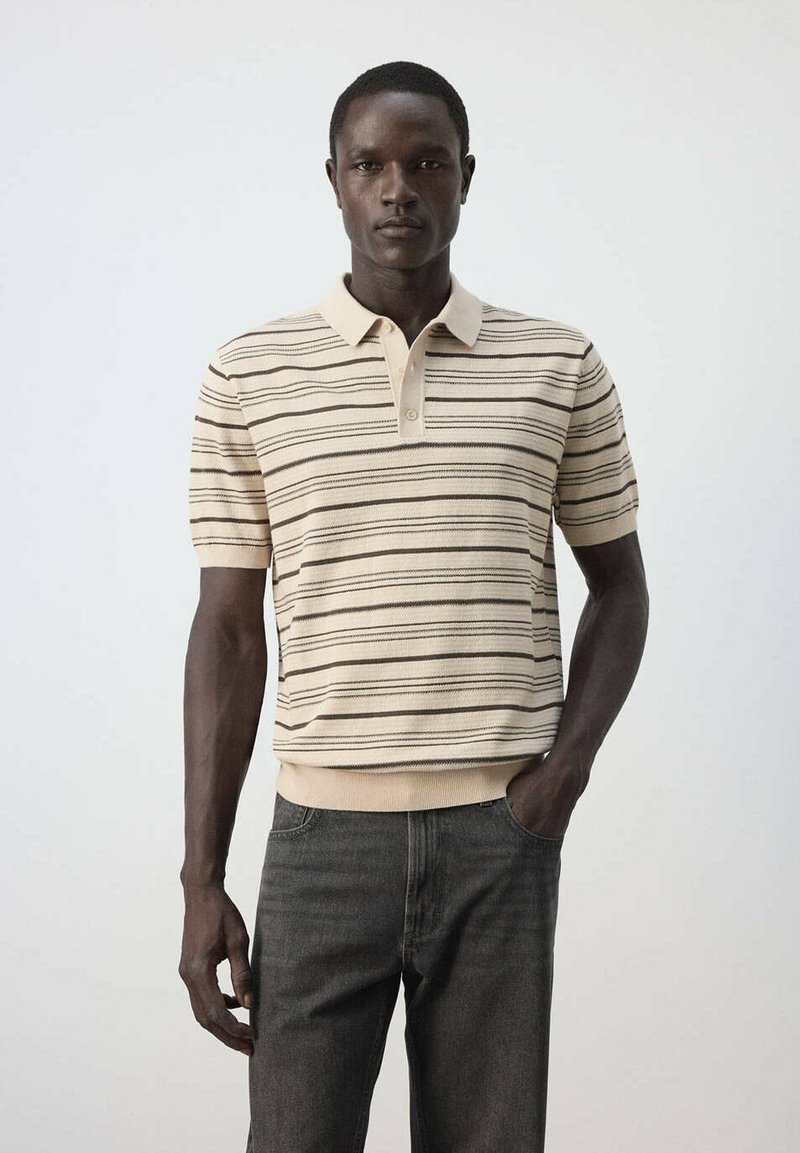 Homme portant un polo beige à manches courtes avec des rayures noires horizontales et un pantalon gris foncé, debout devant un fond uni.