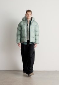 Πρασινο-μίντ μπουφάν με ένθετο και ψηλό γιακά και φερμουάρ μπροστά, φορεμένο πάνω από πουκάμισο καρό και φαρδιά μαύρα παντελόνια, περπατώντας σε ουδέτερο πάτωμα.