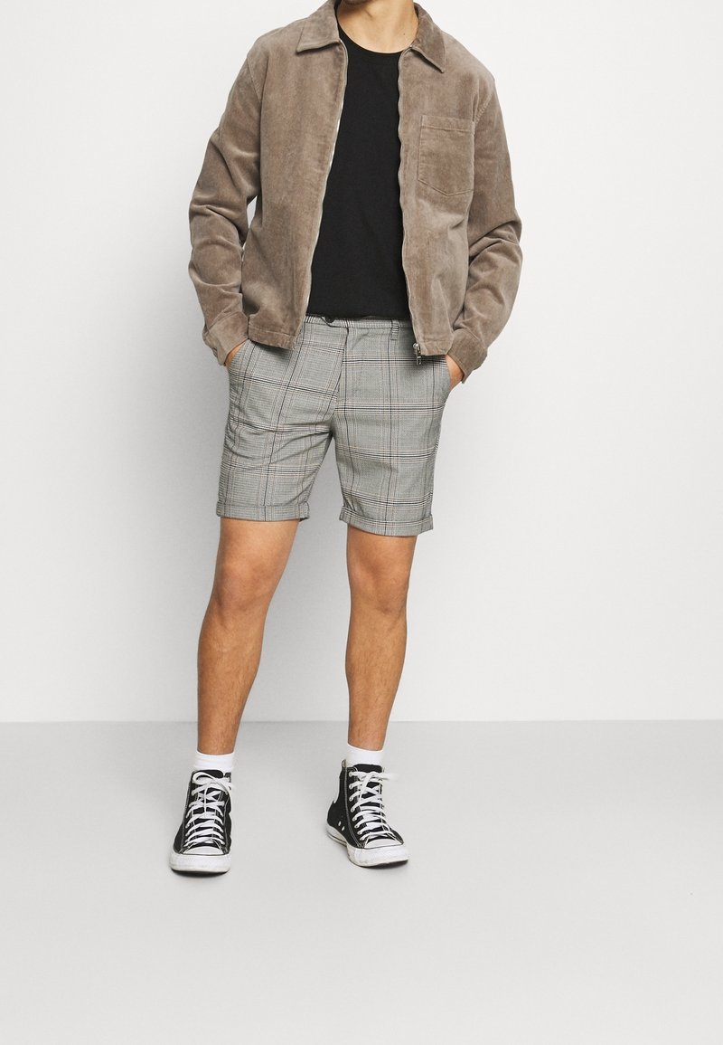 Homme portant une veste marron, une chemise noire, un short à carreaux gris, des chaussettes blanches et des baskets noires montantes, debout les mains dans les poches.