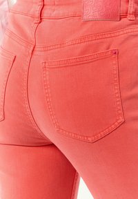 Coral stretch jeans feature a high waist, five pockets, and subtle stitching details. The fabric has a smooth texture and slight sheen.