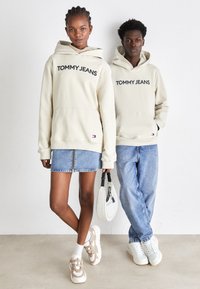Sudaderas beige con el texto "TOMMY JEANS", combinadas con una falda de mezclilla y jeans. Las zapatillas tienen un diseño beige y blanco, con acentos sutiles.