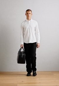 Svart lædertaske holdt af en person i en hvid skjorte og sorte bukser, der står på et trægulv mod en lys grå væg.