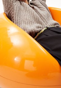 Fauteuil poire en plastique orange à surface lisse ; une personne s'appuie contre son côté, portant un pull gris texturé et une ceinture colorée.