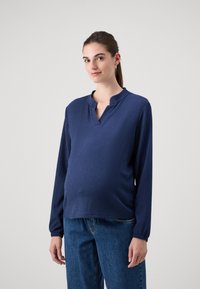 Marineblå langermet graviditetstop med strukturt stoff, featuring en krage med elastisk band og avslappet passform, paret med denimjeans.