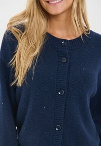 Usmievajúca sa osoba s dlhými blond vlasmi oblečená v tmavomodrom kardigáne na gombíky s jemnými trblietavými detailmi.