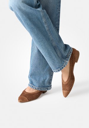 Beine übereinandergeschlagen, blaue Jeans und braune, durchlöcherte Ballerinas mit spitzen Zehen auf weißem Hintergrund.