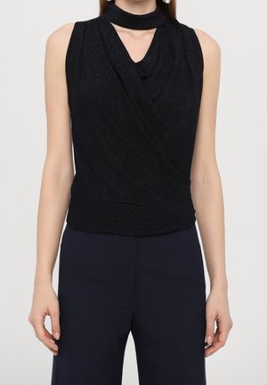 Femme portant un haut noir scintillant sans manches avec un col froncé et un pantalon taille haute foncé, debout devant un fond uni.