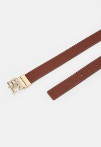 Ruskea nahkavyö, jossa on kultainen metallinen solki, jossa on toisiinsa kiertyvä logomuotoilu; valkoista ommelta reunoilla; säädettävä useilla reijillä.