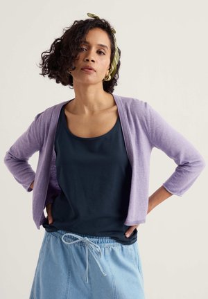 Kvinde med krøllet hår iført en lilla cardigan, sort top, blå bukser med snor og grøn hovedbeklædning, stående med hænderne på hofterne.