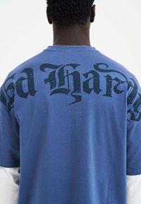 Blaue Baumwoll-T-Shirt mit großen schwarzen gotischen Buchstaben auf dem Rücken. Kurze Ärmel, Rundhalsausschnitt, glatte Textur.