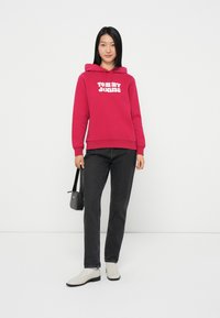 Hoodie fuchsia avec logo "TOMMY JEANS" à l'avant, assorti à un jean noir et des chaussures blanches sans lacets. Modèle tenant un petit sac noir.