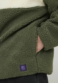 Pull en polaire vert avec bande horizontale crème, surface texturée, poche latérale et patch logo violet en bas. Tissu doux, design décontracté.