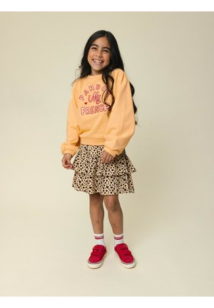 Glimlachend meisje met lang donker haar, gekleed in een gele trui, een rok met luipaardprint, witte sokken en rode sneakers, staand tegen een beige achtergrond.