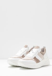 Paire de baskets basses blanches et beiges avec fermeture à lacets et semelles épaisses texturées, posées sur une surface blanche.