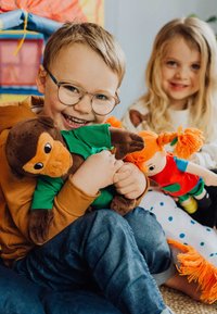 Twee kinderen zitten op een gestreepte kussen; de een houdt een pluchen bruine aap in een groene shirt vast, de ander knuffelt een pop met oranje vlechten.