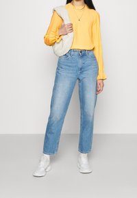 Top amarillo de manga larga con puños fruncidos, combinado con jeans de cintura alta de color azul claro y zapatillas blancas; llevando un bolso de color crema y peludo.