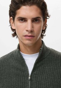 Maglione a coste verde scuro con collo a zip e cerniera argentata, indossato sopra una camicia bianca, con tessuto texturizzato e design aderente.