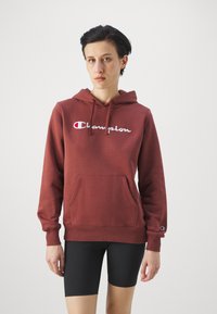 Bordó kapucnis pulóver, elülső zsebbel, húzózsinórral és hímzett "Champion" logóval. Sima anyag és bordázott mandzsetta. Fekete rövidnadrággal viselve.