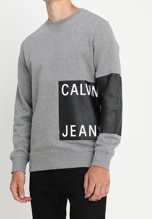 Mężczyzna w szarej bluzie z czarną naszywką na przodzie i prawym rękawie z napisem „CALVIN JEANS".