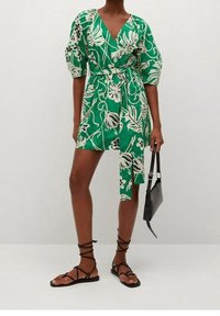 Robe portefeuille verte à motif floral avec manches bouffantes, décolleté en V et taille nouée. Portée avec des sandales plates noires et un petit sac à main noir.